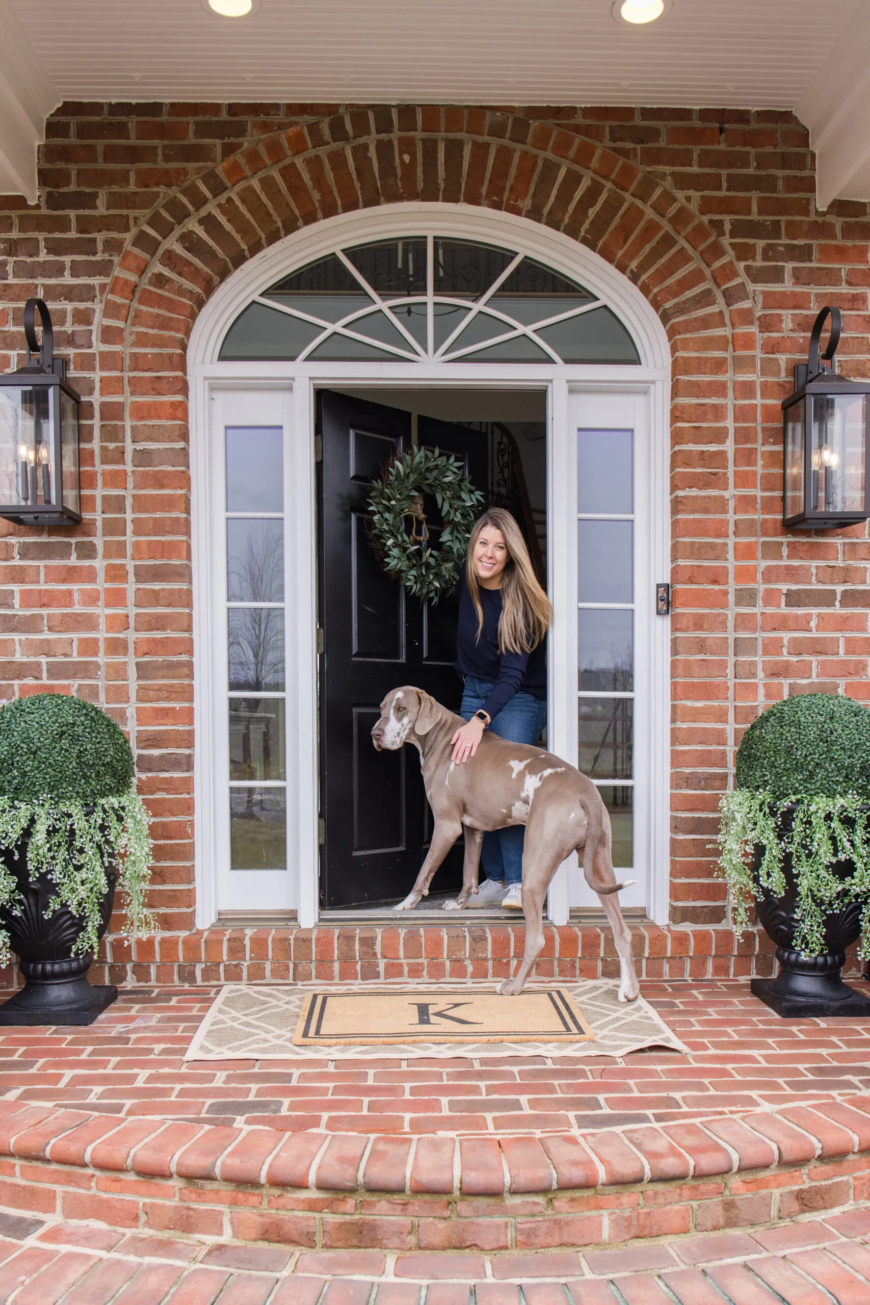 Spring front porch styling Arched Manor spring front porch styling featuring Megan and dog Slinky | inital doormat | outdoor rug | large shrubs styling in black planter with hanging plants | black wall sconce | olive wreath on front door