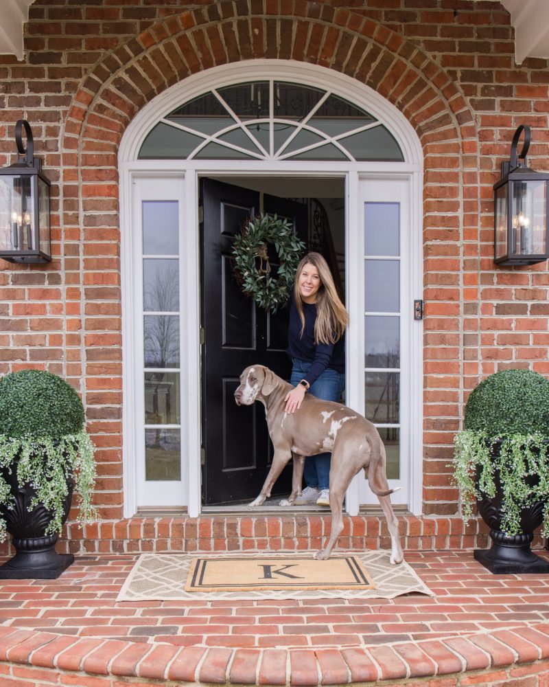 Spring front porch styling Arched Manor spring front porch styling featuring Megan and dog Slinky | inital doormat | outdoor rug | large shrubs styling in black planter with hanging plants | black wall sconce | olive wreath on front door
